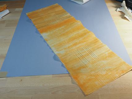 The first part of the folding process is precreasing the grid necessary to make all the folds.<br>Here, the vertical precreases are already folded (compare the length of the sheet to the previous photo).